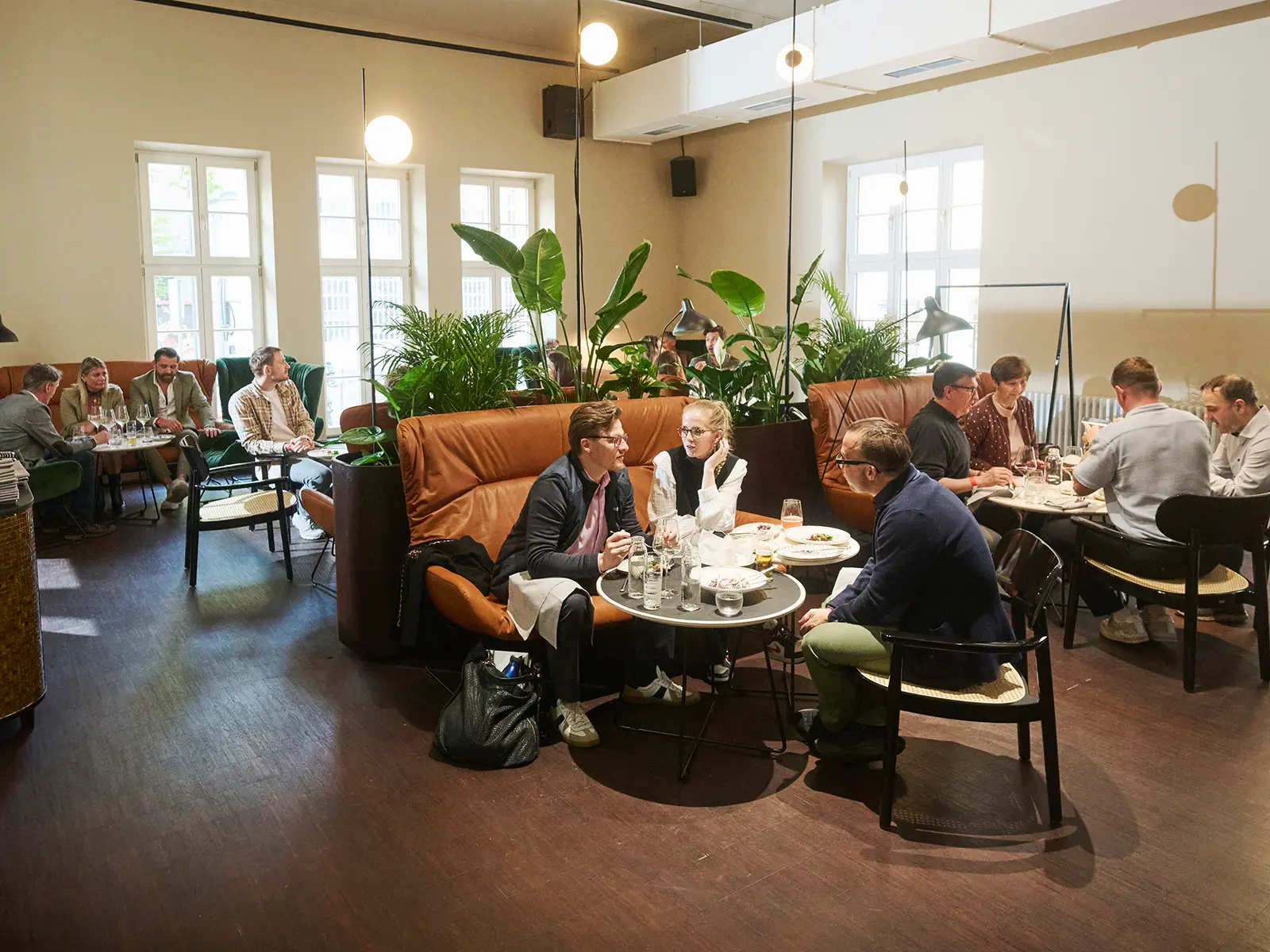 Gäste an Tischen in der Bel Etage des sophie7, mit Ledersofas, Pflanzen und hohen Fenstern