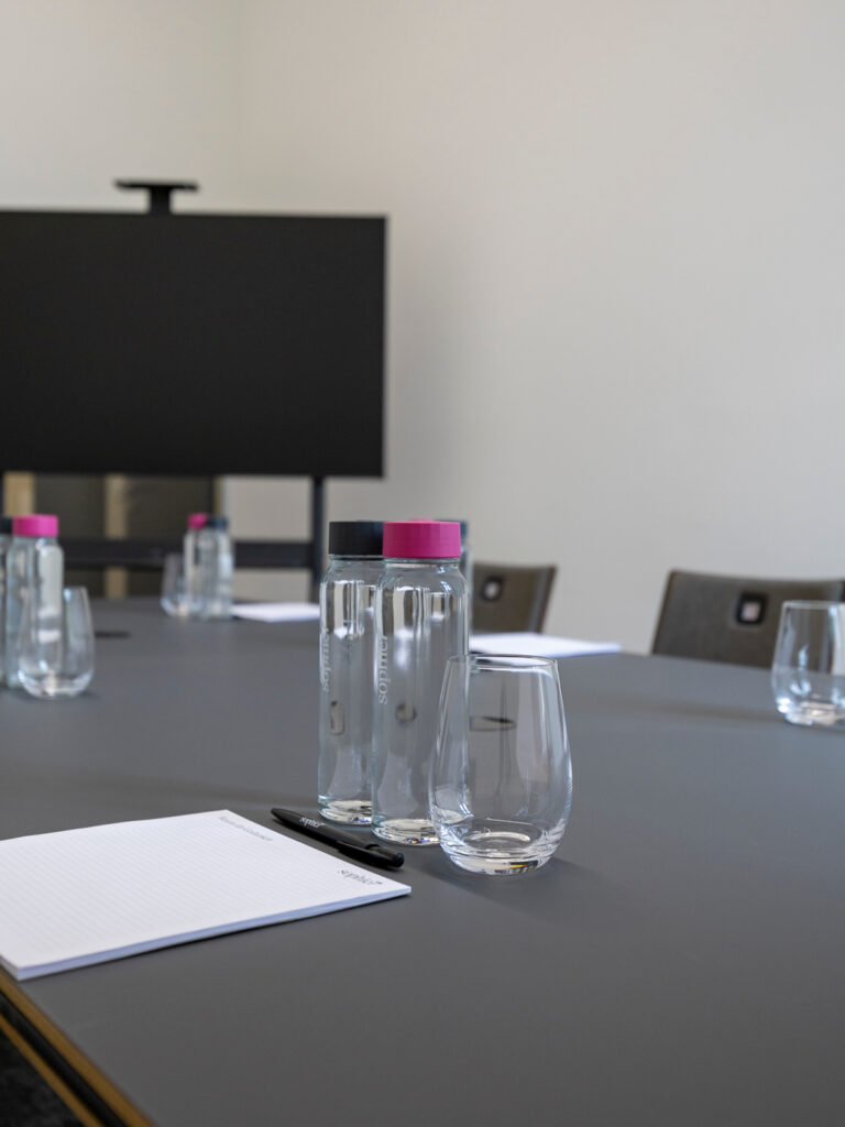 Gedeckter Meetingtisch mit sophie7-Wasserflaschen, Gläsern, Notizblock und Bildschirm