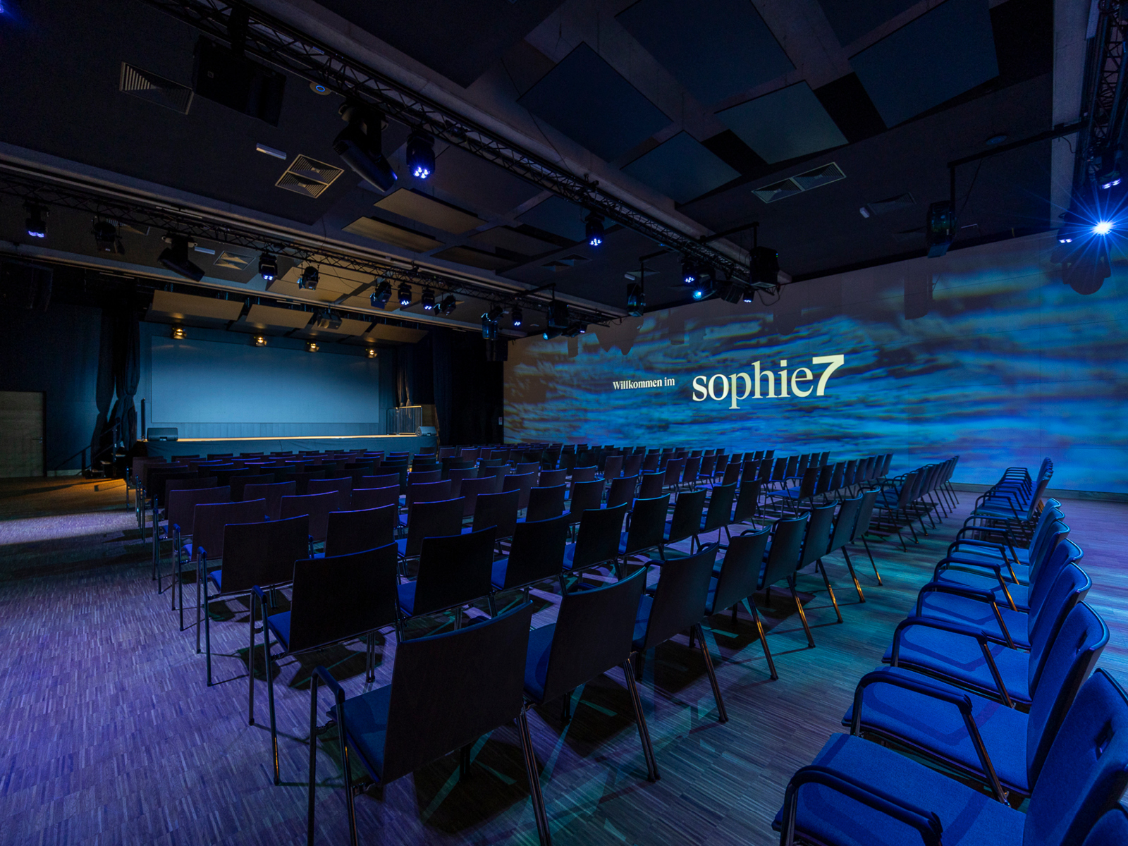 Großer Eventsaal mit Reihenbestuhlung und sophie7-Projektion in blauem Licht