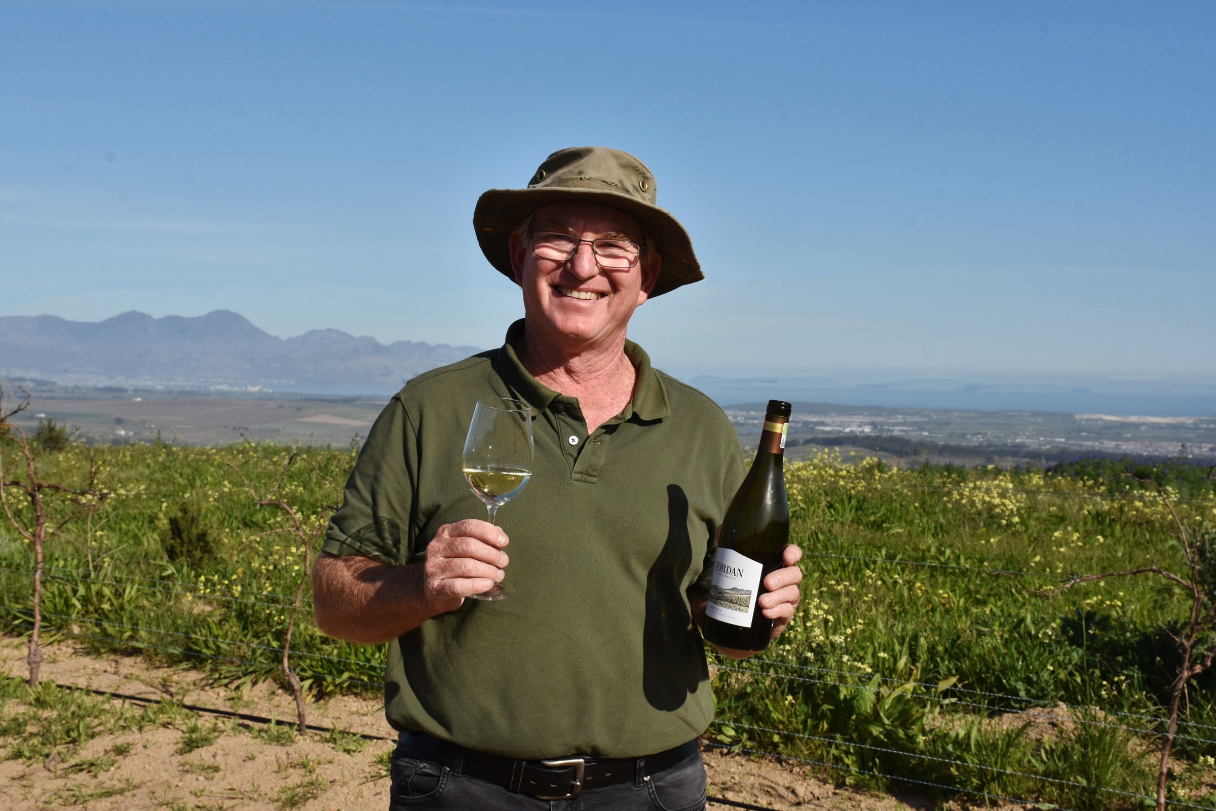 Winzer Gary Jordan mit Weinglas und Flasche im Weingarten, im Hintergrund die Berge von Stellenbosch